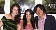 The heads and upper bodies of three people side by side, posing for the camera smiling. At the left, and centre are two white women in their mid-twenties, both with long dark-brown hair. The woman on the left is wearing black and green clothing, and the woman in the centre is wearing pink. At the right is a Japanese man in his early-thirties, with dark centre-parted hair. He is wearing a pink open-collar shirt, and a black suit-jacket.