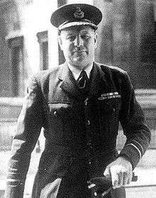 Half-portrait of man in dark military uniform with peaked cap, carrying gloves in left hand