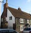 Three-quarter view of an end-of-terrace two-storey cottage with a tiled roof, side chimney-stack, irregular cobblestone façade (lit by sunlight) and wide windows with red-brick surrounds. Parked cars partly obscure the view.