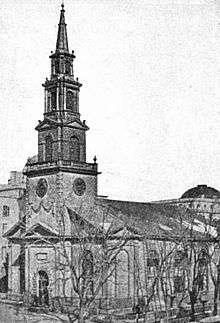 A black-and-white photograph of a church with a tall steeple, seen from its left across the street