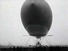 An airship is seen with its pointed bow towards the camera; the gondola is suspended below the envelope with a stationary propeller on each side. level with the bottom of the envelope are two horizontal ailerons