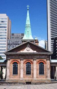 Photo showing of the end of the church. It is of brick and has three round-headed windows set between four sandstone pilasters, surmounted by a Classical lintel and pediment. At the further end of the church is a spire sheathed in pale green copper.