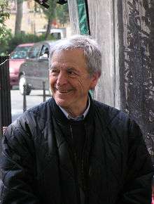 Photograph of the head and upper body of a smiling man looking well to the left of the camera. He is wearing a collared shirt and a pullover sweater underneath a jacket. He is apparently sitting, and well behind him there is a street with two cars visible.