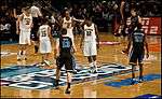 Two opposing basketball teams on a basketball court. One team is in white uniforms with the word Michigan on the front and names on the back and the other is dark blue with the word Duke on the front and names on the back.