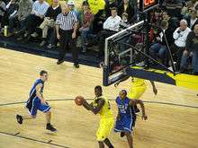 A basketball player in a maize uniform with his back to the basket maneuvers with the basketball. Defenders in light blue uniforms attempt to contest him.