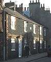 A row of short cottages with four evenly-spaced doors opening on to a low pavement. Beyond the furthest cottage is a taller, blank-walled house with a wide, tall chimney, and six other chimney-pots are at least partly visible. The cottages have identical dark cobblestone walls and red-brick dressings around the windows and between the lower and upper storeys.