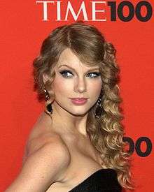 Taylor Swift stands in a Time press area, wearing a black, strapless dress and curled hair