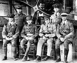 Six men in military uniforms with peaked caps, two standing and four seated, in front of a biplane