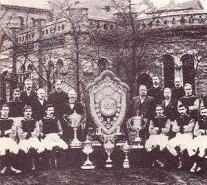 A sepia photograph with a large old structure in the background obscured by trees. In the foreground there is a large shield surrounded by five trophies. On either side of the shield stands 8 people.