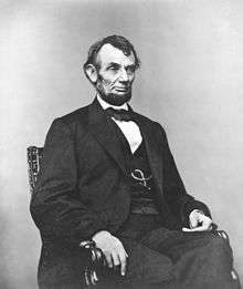 Middle-aged man in a beard posed sitting in a suit, vest and bowtie.