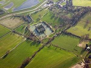 An aerial view of the Bodiam. The square castle is surrounded by a roughly square moat, and beyond that stretch green fields.