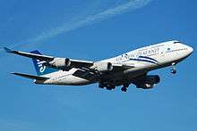  An Air New Zealand 747-400 with its landing gear down and flaps down.  The aircraft is mostly white and blue aircraft and in-flight against a blue sky. On each of the two wings are two engines.