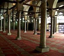 A large room filled with rows of cylindrical columns on top of square bases. The columns support arches which are pierced by square beams going the length of the room in both directions. Hanging from the beams are lamps, and the room's floor is covered with a red carpet with a repeated beige arched doorway shaped design on it. Exterior light enters from right of the room.