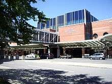 Three-story brick building with blue windows; a glass pedestrian bridge travels from building to unseen parking garage on left, crossing entrance road. "ALBANY INTERNATIONAL AIRPORT" sign is visible on side.