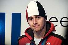 A brown-haired and brown-eyed young man, with a goatee, wears a red-white-and-black winter cap, and a similarly colored black-lined hooded jacket over a white t-shirt.