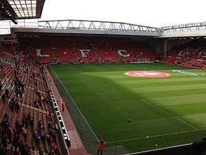 A two-tiered stand which has red seats, there are also white seats which spell out "L.F.C.. In front of the stand is a field of grass