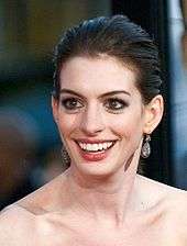 A bust shot of a young woman standing in a side view, her head turned to look to the camera. Her long hair is pulled back from her face and cascades down her back. She wears a black sleeveless dress with a gold trim around the back and under her arm. She wears jeweled silver floral shaped earrings and smiles softly. There is a crowd of people, slightly out of focus, in the background.