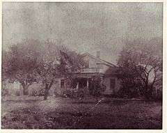 black-and-white image of a clapboard,  1 1⁄2-story, pitched-roof house, looking toward house through several trees from across yard