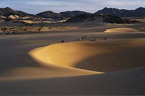 A picture of a large, depressed area of sand to the right and a mountain range in the background. Relatively small vehicles are parked alongside the massive hole.