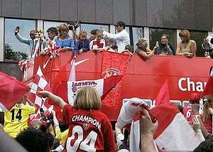 A group of people on a red open-topped bus wave to a crowd of onlookers.