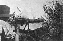 A soldier lies amongst the grass in the prone position behind a machine gun which he is holding in the shoulder.