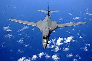 Top view of B-1B in-flight with white clouds scattered underneath. Its wings are swept fully forward as the gray aircraft flies over the ocean.