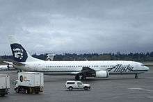 Right side view of an airplane taxiing on the tarmac, with several trucks in the foreground and to the left. In the background is a tree-covered hill and dark clouds.