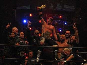 Ten members of the Bullet Club group celebrate in a professional wrestling ring. Several are using a hand gesture, extending the index and little finger, with the tips of the renaming fingers touching