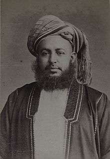 A black-and-white photograph of a man with a dark beard wearing a turban, a dark jacket, a shirt, and a belt, sitting in a chair, and looking at the viewer