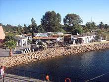 Colour photo showing the reconstruction of the ancient port of Palos de la frontera: transportation barrels, low buildings bordered by a covered gallery, a chariot at the very edge of the water awaiting the arrival of a boat.