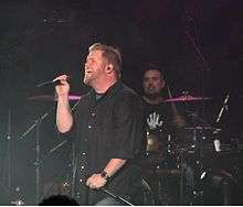 In a dark room, a singer holding a microphone in one hand and stand in the other is standing in front of a drummer