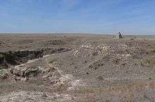 Dry grassland with a small rocky canyon; pyramidal stone monument on hill nearby