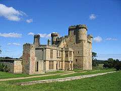 A large stone built ruined building. The nearer part is two stories with square windows; behind is a tall square keep with turrets and battlements. In the foreground is grass with a low stone wall; in the background a blue sky with a few white clouds.