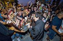 A crowd of people is all looking towards a man at the center who is signing a hat. The crowd is attempting to hand him posters to sign and others are taking pictures using cameras and cell phones.