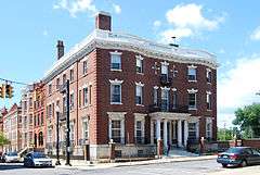 A three-story brick building on a street corner seen from across the intersection. It has an entrance with columns and a white balustrade on its flat roof.