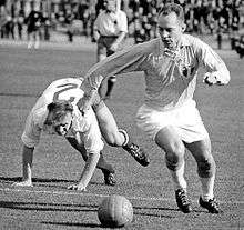 A black-and-white action shot taken in the midst of an association football match. To the viewer's right a player in light shirt, white shorts and white socks turns to his right, his eyes pointed down towards the ball. A shield bearing the letters "MFF" is prominently displayed on his shirt. Behind him, an opposing player in a white shirt can be seen stumbling mid-chase, throwing his arms forward to break his fall.