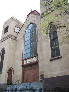 Most of the front of a three-story building is visible. It shows two rectangular towers, one on each side of a recessed bay, all clad in tan stucco. The towers have pointed arched windows on the bottom and square ones on top. The bay has four wooden doors at the bottom and a sign with Hebrew writing on top of them, surmounted by large arched multi-paned window. Atop the roof of the bay is a small metal Star of David.