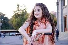 A young girl with long wavy light brown hair, pale skin and deep brown eyes wearing an orange dress sits on a bicycle and looks off into the distance.