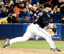 A man in a black baseball jersey, cap, and white pants pitches a baseball with his left hand.