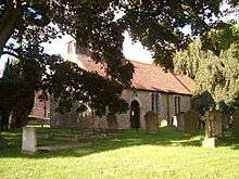 A stone church with a red tiled roof seen between trees in a churchyard. On the west gable is a bellcote.