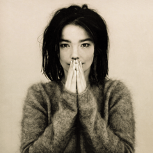 A picture of the album cover depicting a muted background with Björk standing facing forward in the middle. Björk is dressed in a fuzzy ragged sweater with her hands close together covering most of her mouth.