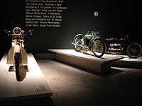 Three pristine 1920s - 1930s motorcycles on pedestals in front of text printed on the wall with words relevant to the period, such as "Prohibition" and "Al Jolson".