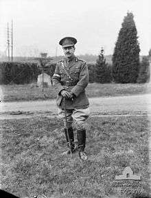 A man in a neat military uniform with a peaked cap, ribbons on his tunic, Sam Brown belt, cane, riding boots and spurs.