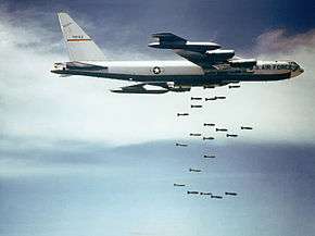 Against a blue sky with white clouds, a B-52F releases bombs over Vietnam.