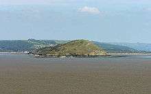 Green covered rocky land in expanse of sea. Hills behind.