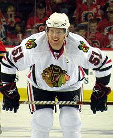 Hockey player in white uniform with a picture of crossed hockey sticks on the shoulder and the painted face of an indigenous person on the chest. He crouches and holds his stick on his knees, smiling.