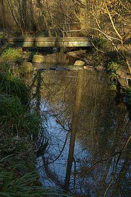 A stream in the foreground crossed by a concrete footbridge, shaded by trees.