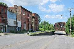 Several old brick buildings along the left side of a street, which descends underneath a rusted metal bridge.