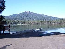 A parking lot with a forest-lined lake behind it, and a gentle sloping mountain in the background