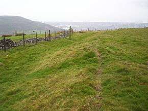 A grass-covered bank and ditch run alongside a dry-stone wall.